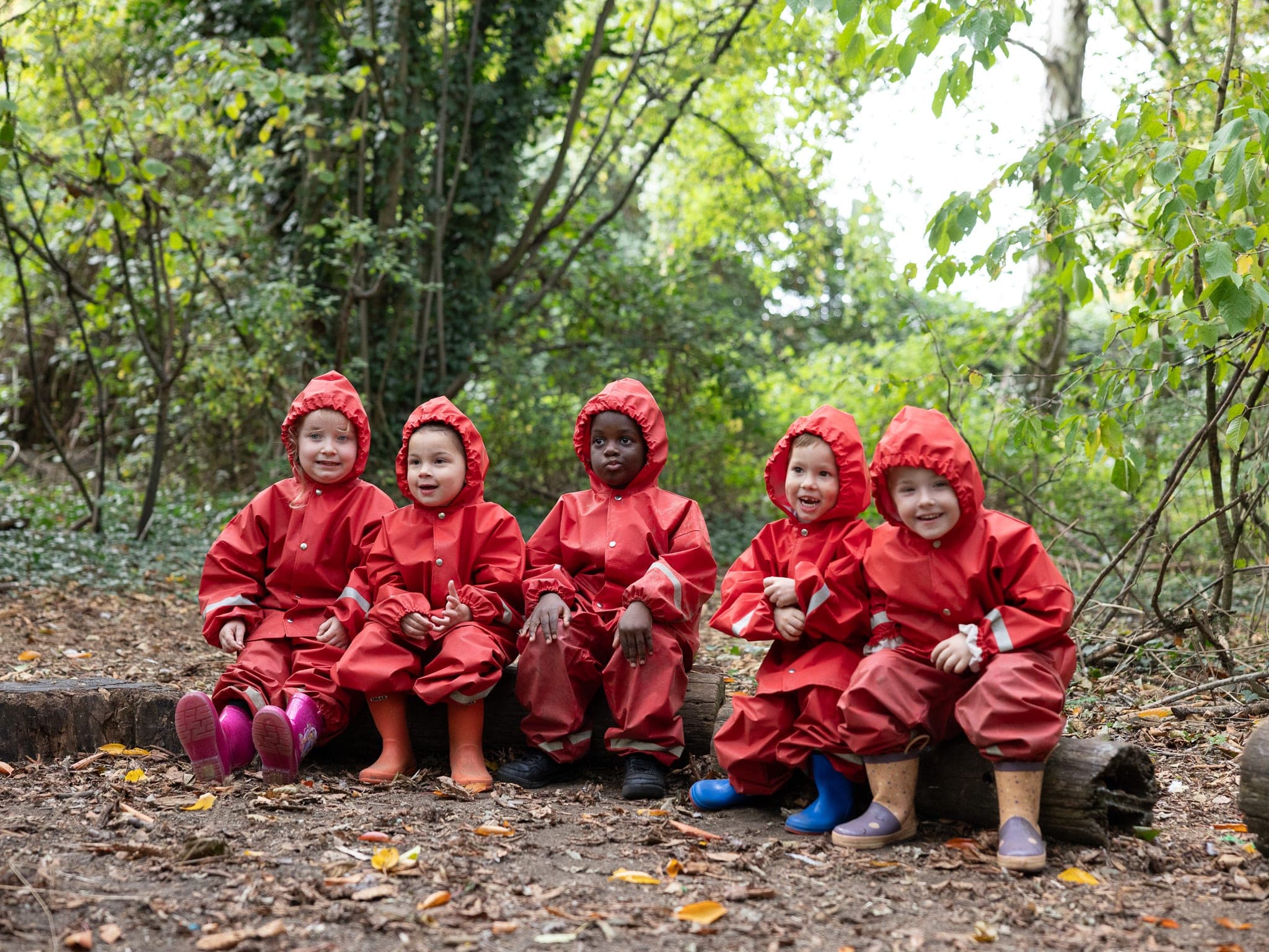 Young children in red waterproof suits sitting on a log in the woods during forest play