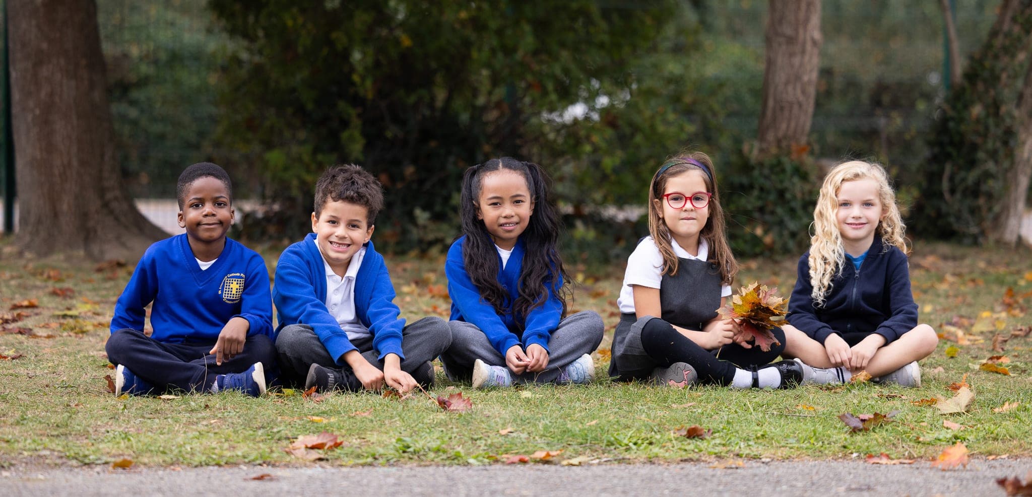 Five pupils in blue jumpers sitting together on the school field, smiling