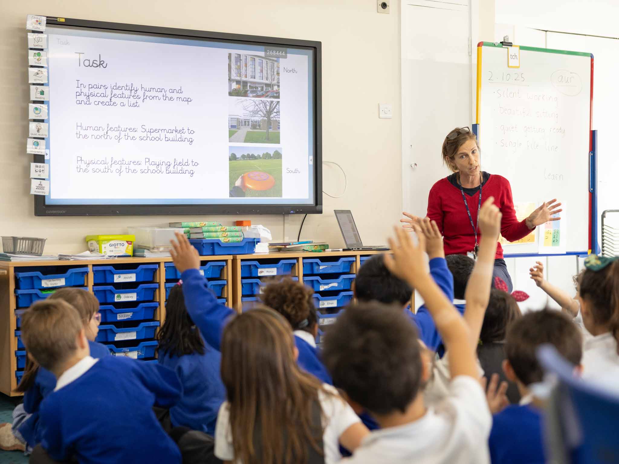 Pupils raising their hands on the carpet as their teacher leads a lesson
