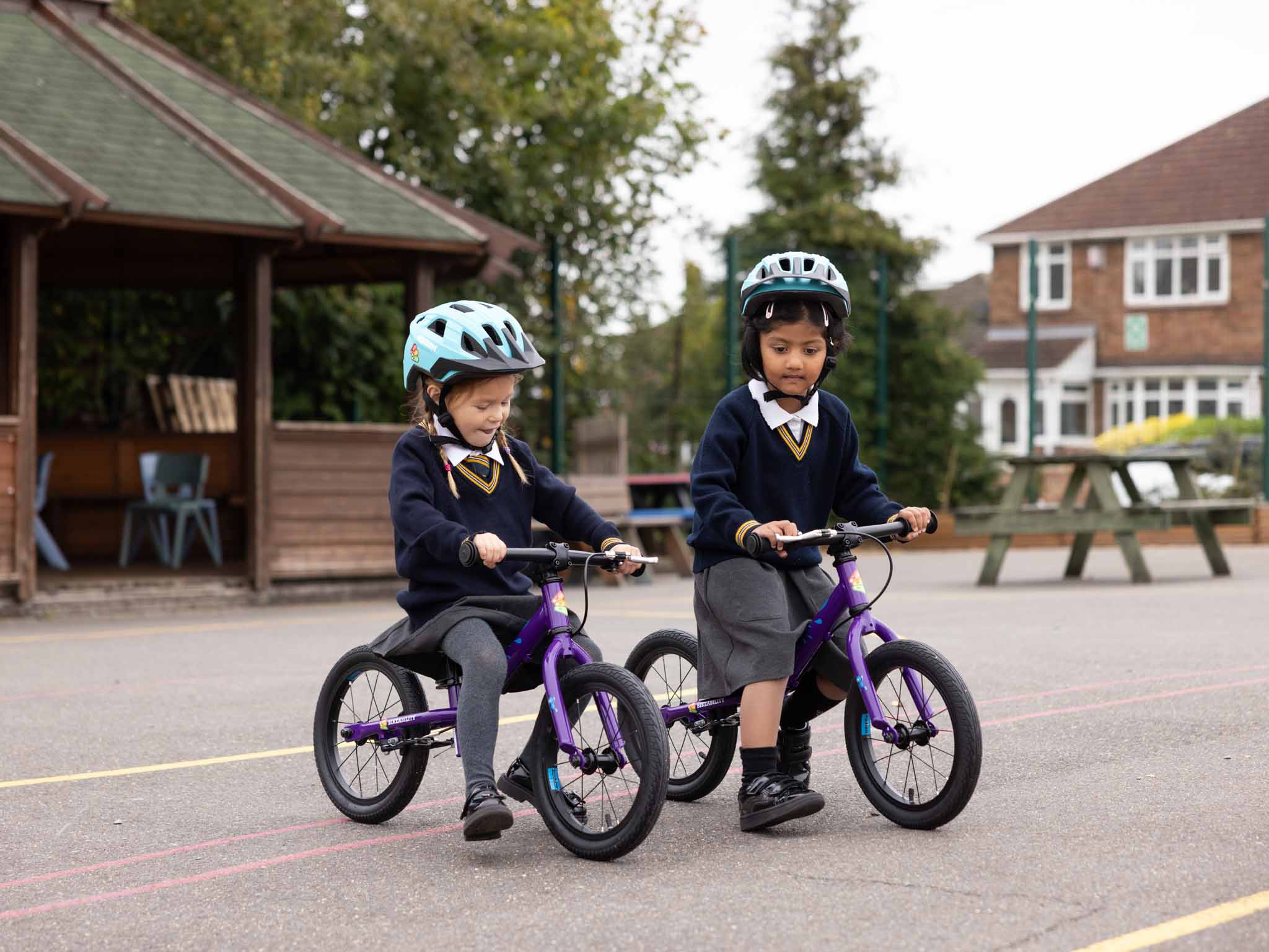 Two young children in helmets riding balance bikes in the playground