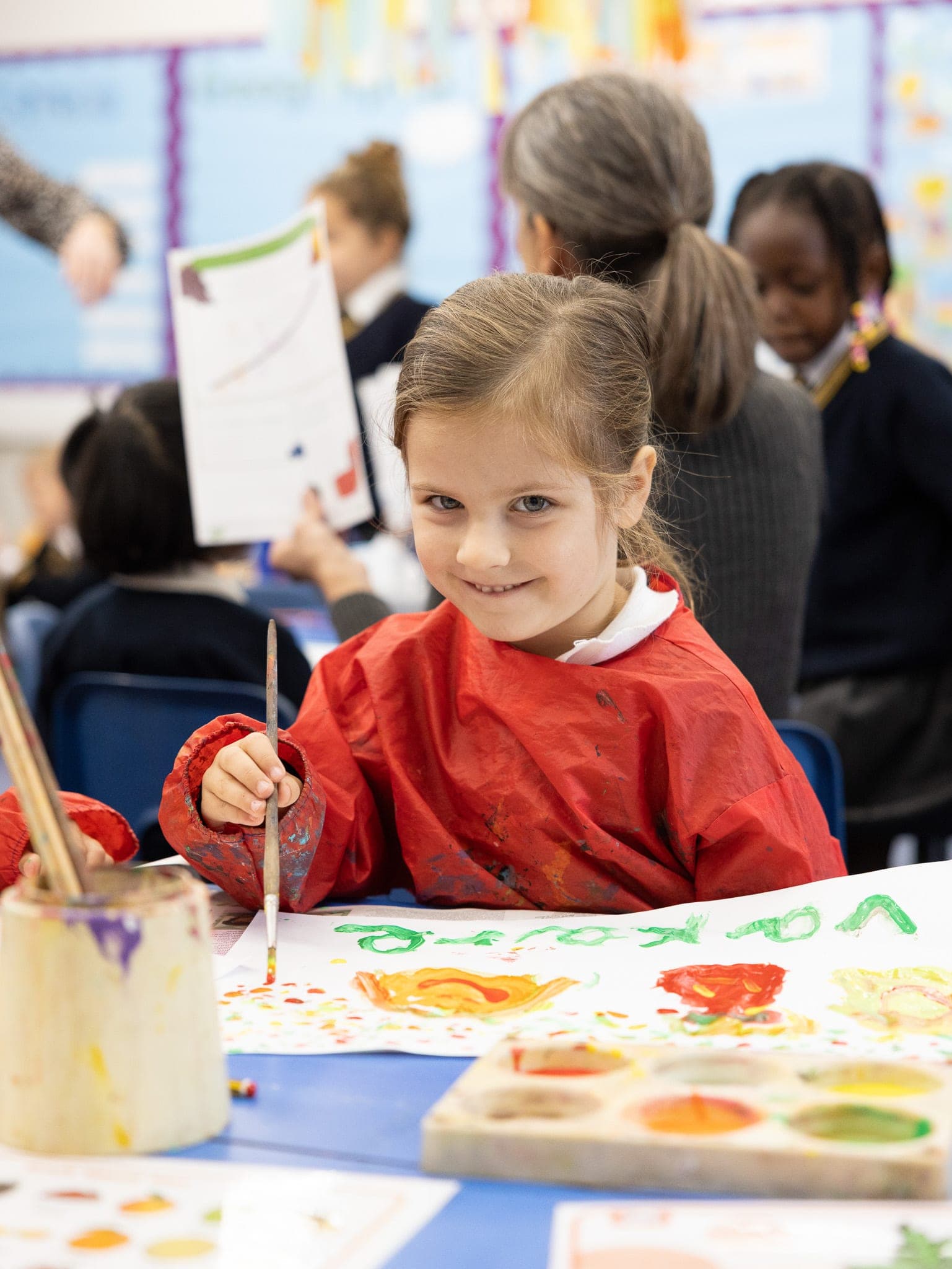 A smiling girl in a red apron painting at her table