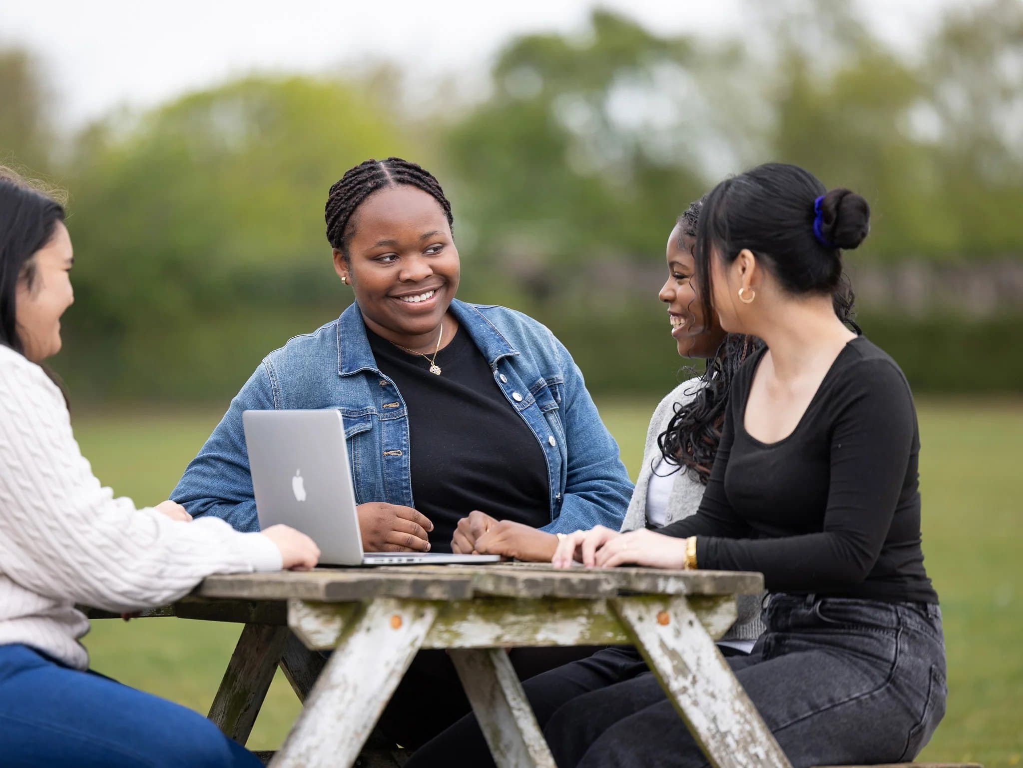 Sixth form students laughing together around a laptop at a picnic bench on the field