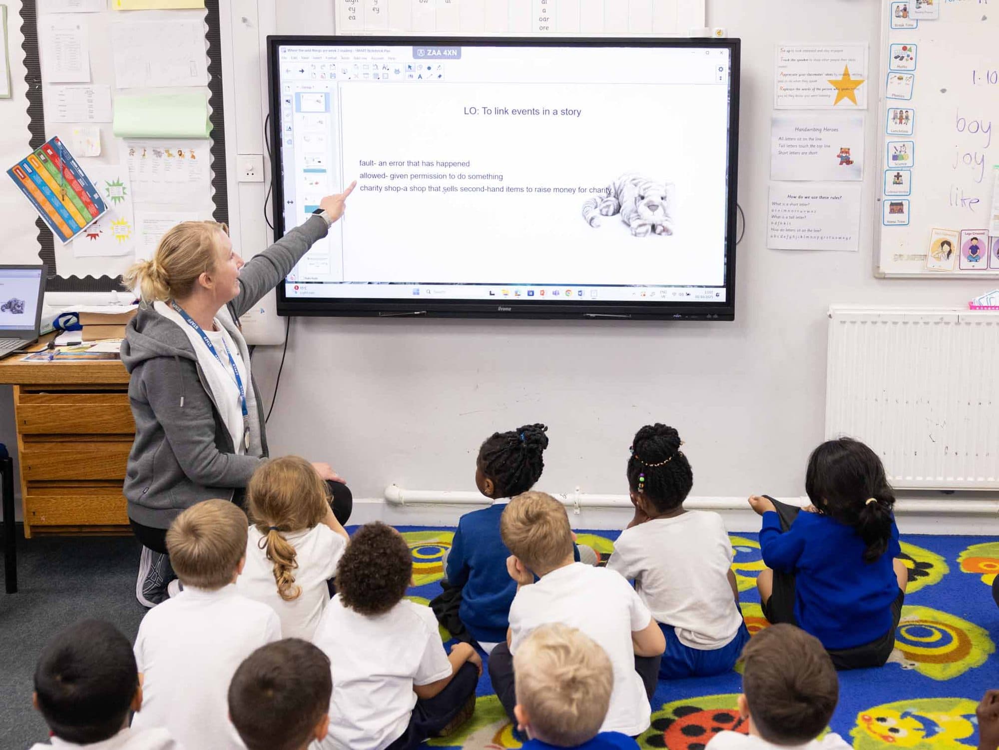 Infant class sitting on the carpet as their teacher points to the screen