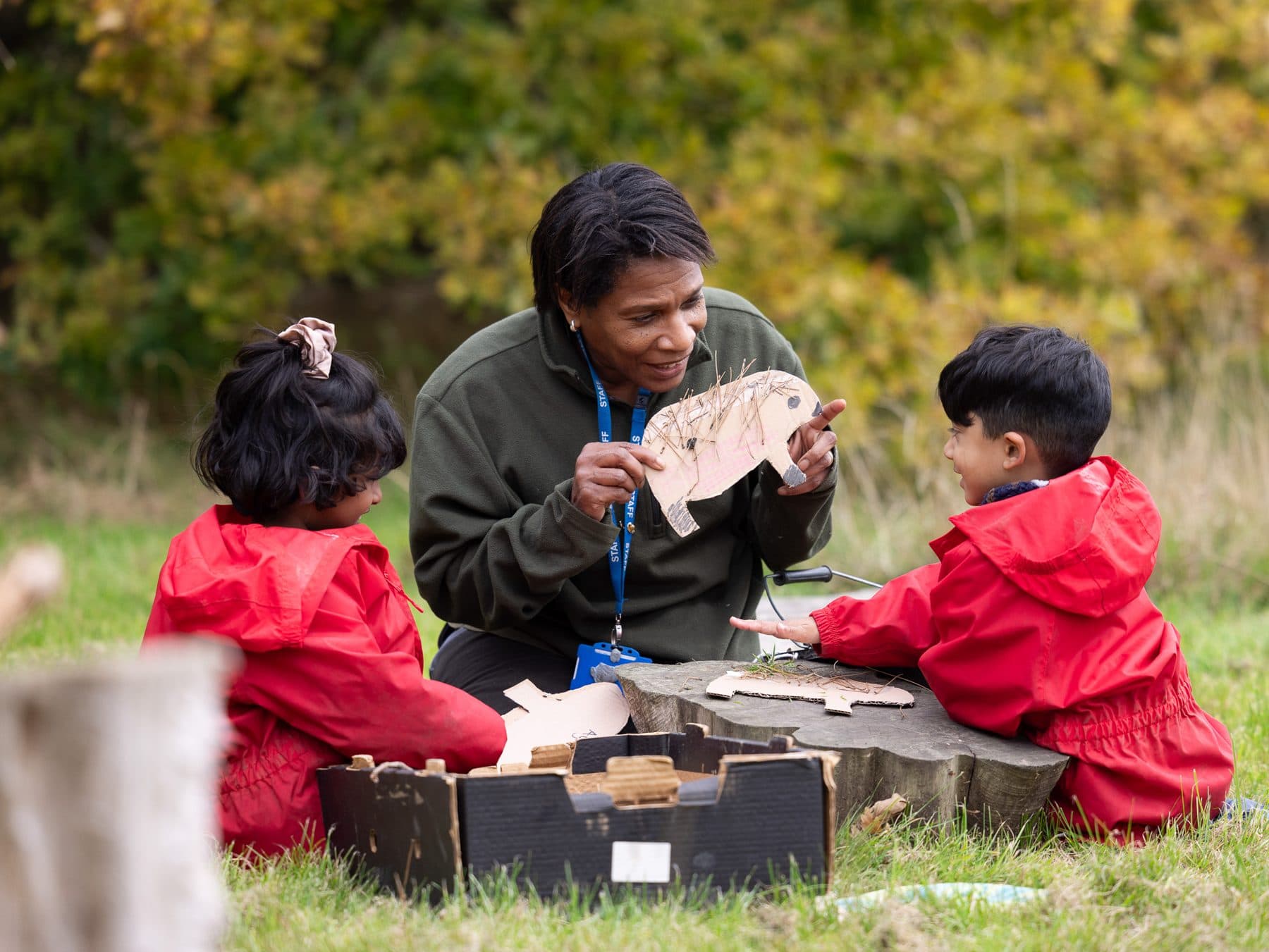 Teacher and two young children in red coats with a wooden hedgehog in the school grounds